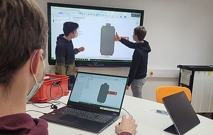 Ein Schüler am Tablet verfolgt, was die anderen beiden am Whiteboard zeigen; auf den Bildschrirmen das Modell eines Desinfektionsmittelbehälters