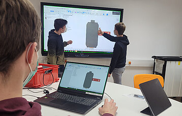 Ein Schüler am Tablet verfolgt, was die anderen beiden am Whiteboard zeigen; auf den Bildschrirmen das Modell eines Desinfektionsmittelbehälters