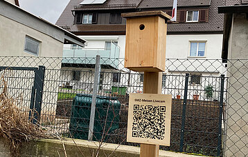Meisenkobel und QR-Code zum Projekt des Stadtmedienzentrums Stuttgart auf dem Parkplatz, hinten ein Haus