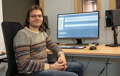 Dejan Simonović, der Leiter der ComputerSpielSchule Stuttgart sitzt auf einem Stuhl an einem Schreibtisch und lacht in die Kamera. Im Hintergrund sieht man einen großen Bildschirm.