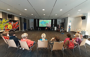 Stuhlreihe mit Kindern von hinten im Filmsaal auf Projektionswand blickend